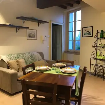 Accogliente spazio open space rustico che unisce una zona pranzo con tavolo in legno apparecchiato e un soggiorno con divano confortevole sotto travi a vista. Vista su finestra e mobile in ferro battuto. Catina Basi