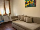 Confortevole soggiorno moderno con un grande divano grigio, pavimento in legno lucido e un vivace dipinto astratto sulla parete. Una sedia blu &egrave; vicina alla finestra. Catina Basi