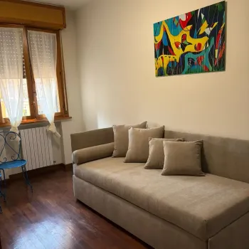 Confortevole soggiorno moderno con un grande divano grigio, pavimento in legno lucido e un vivace dipinto astratto sulla parete. Una sedia blu &egrave; vicina alla finestra. Catina Basi