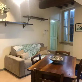 Accogliente zona giorno open-space con soffitto a travi a vista, tavolo da pranzo in legno massiccio, divano invitante e scaffali decorativi. Stile rustico e confortevole. Catina Basi