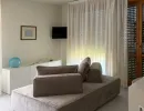 Soggiorno moderno e luminoso con un divano modulare grigio vellutato. Dispone di una console bianca con un accento decorativo blu e una TV a parete. Le tende trasparenti si affacciano su un balcone. Catina Basi