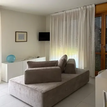 Soggiorno moderno e luminoso con un divano modulare grigio vellutato. Dispone di una console bianca con un accento decorativo blu e una TV a parete. Le tende trasparenti si affacciano su un balcone. Catina Basi
