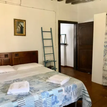 Camera da letto rustica con travi a vista e pavimento in cotto. Presenta un letto con testiera in legno, copriletto azzurro e una scala a pioli decorativa. Catina Basi
