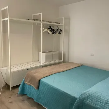 Camera da letto moderna e luminosa dal design minimalista. Presenta un letto matrimoniale con copriletto azzurro e un pratico guardaroba aperto bianco. Catina Basi